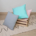 Poszewka jersey 40x40 w kolorze antracytowym leży na jasnym dywaniku, obok znajdują się poduszki w kolorach błękitnym i różowym, umieszczone w drewnianym koszu. Tło jest neutralne, a materiał poszewki jest gładki i teksturowany.