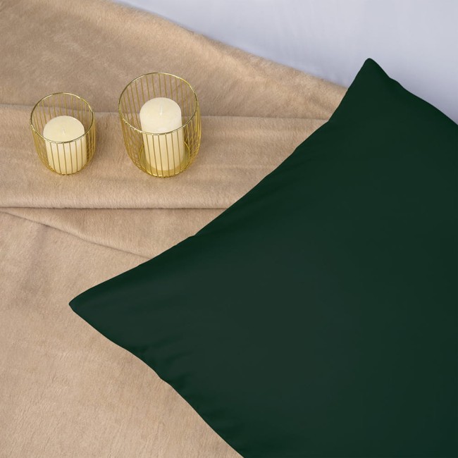 Poszewka satynowa 50x60 w kolorze butelkowej zieleni, leżąca na beżowym tle obok dwóch świec w złotych osłonkach. Zbliżenie ukazuje gładką fakturę materiału, która podkreśla jej elegancki charakter.