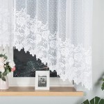 Firana gotowa Demetria żakardowa w kolorze białym o wymiarach 300x160 cm, zawieszona w oknie. Wzór z delikatnymi kwiatami i ażurowym wykończeniem u dołu. Produkt w świetle, z boku widocznego ramka ze zdjęciem i kwiaty na parapecie.
