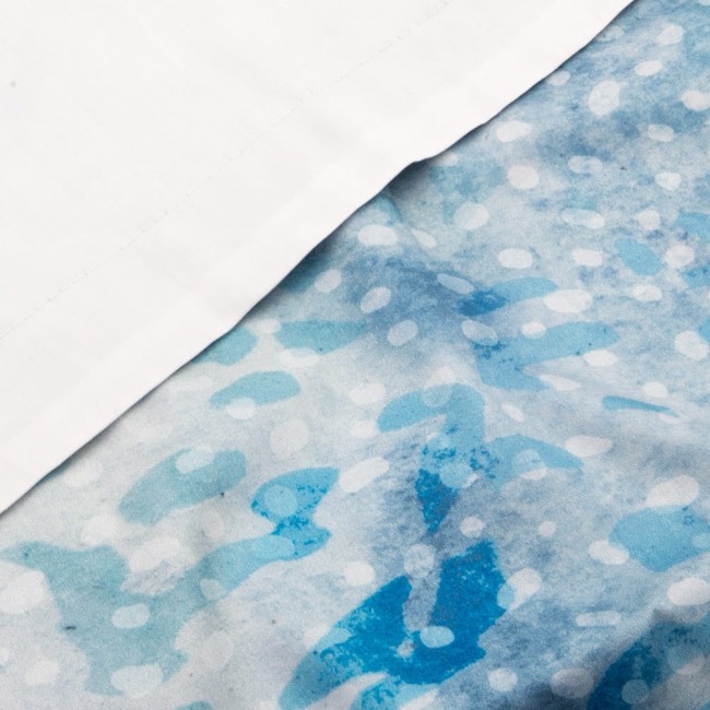 Pościel satyna bawełniana 160x200 Luxury Premium Belize w kolorze niebieskim i białym, z delikatnym, abstrakcyjnym wzorem w odcieniach błękitu. Widoczne białe obszycie. Produkt leży, ukazując szczegóły tkaniny i układ wzorów.