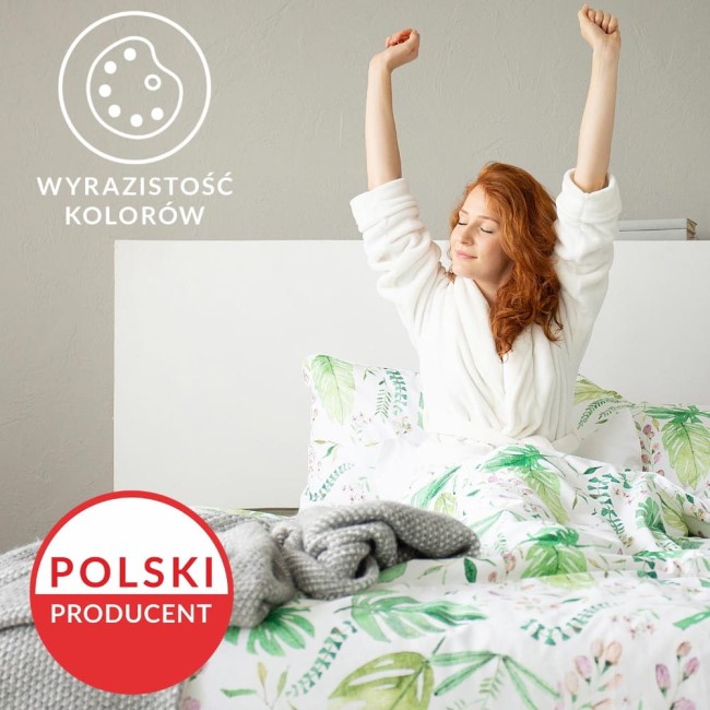 Pościel bawełniana 220x200 cm w motyw wiosennych różowych kwiatów i zielonych liści, rozłożona na łóżku. Widać poduszkę i narzutę, a na wierzchu leży szary, pleciony koc. Tło w jasnym odcieniu szarości, prezentująca przyjemne wrażenie komfortu.