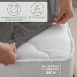 Prześcieradło jersey z elastanem w kolorze antracytowym, rozłożone na materacu o grubości 25 cm, trzymane przez rękę. Widać elastyczny brzeg z gumką. Widoczne napisy: bardzo elastyczne i rozciągliwe oraz wytrzymałe. Tło neutralne.