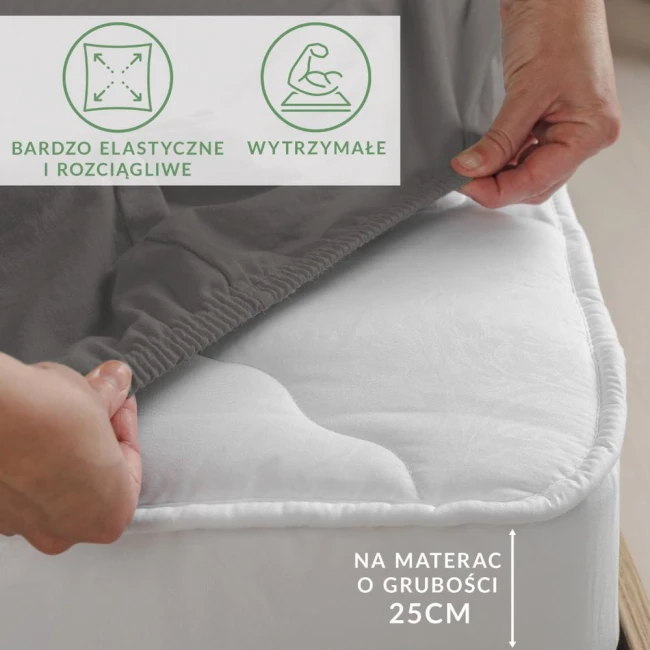 Prześcieradło jersey z elastanem w kolorze antracytowym, rozłożone na materacu o grubości 25 cm, trzymane przez rękę. Widać elastyczny brzeg z gumką. Widoczne napisy: bardzo elastyczne i rozciągliwe oraz wytrzymałe. Tło neutralne.