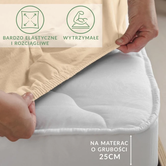 Beżowe prześcieradło jersey z elastanem, rozłożone na materacu, z elastyczną gumką na brzegach. Widać rękę chwytającą materiał, co podkreśla jego elastyczność. Informacje o przeznaczeniu dla materaca o grubości 25 cm. Tło neutralne, materiał gładki.