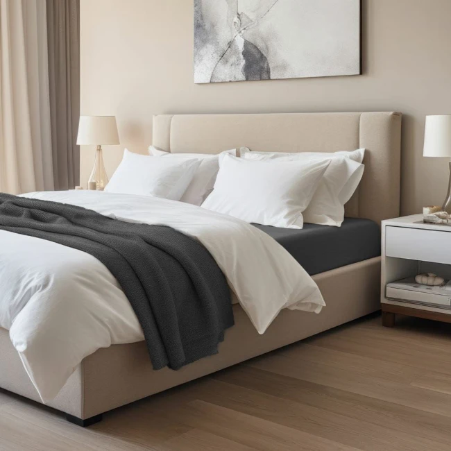 Prześcieradło jersey z elastanem 90x200 w kolorze czarnym, rozłożone na materacu pod białymi poduszkami. Produkt leży na łóżku w jasnym wnętrzu, obok widoczna narzuta w szarym kolorze oraz lampka nocna. Tło w neutralnych odcieniach.