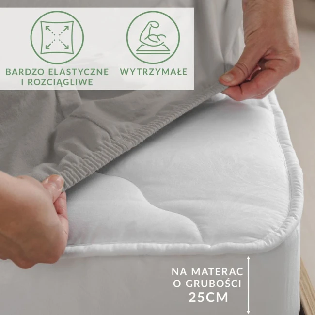 Prześcieradło jersey z elastanem w kolorze popielatym, elastyczne, z gumką, częściowo rozłożone na materacu o grubości 25 cm. Widoczna dłoń zakładająca prześcieradło, tło neutralne, bez wzorów czy dekoracji.