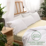Prześcieradło jersey z gumką 70x140 w kolorze kremowym, leżące na materacu, obok białych poduszek. Na tkaninie widoczna grafika informująca o idealnym dopasowaniu do materaca. Tło lekko zaaranżowane z roślinnością i naturalnym światłem.