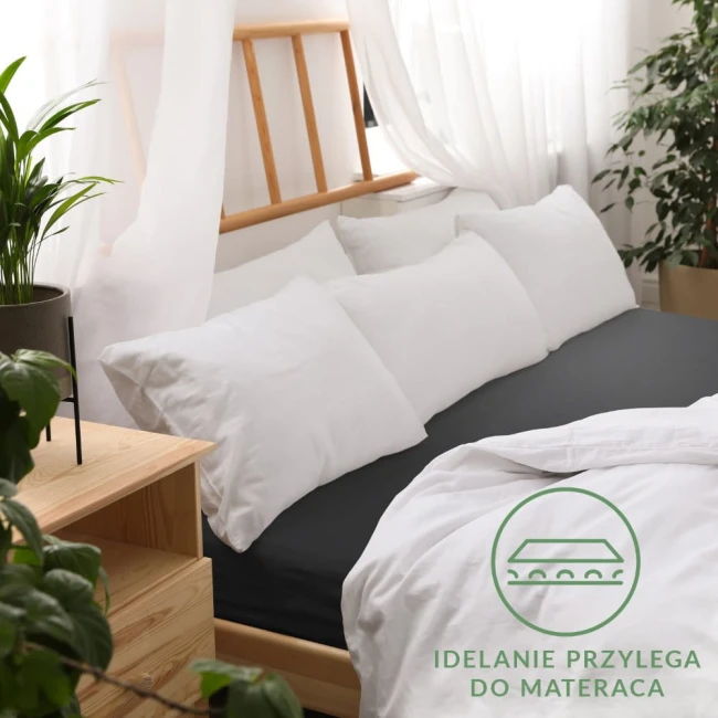 Prześcieradło jersey z gumką w kolorze czarnym, rozłożone na łóżku o wymiarach 120x200 cm. W tle widoczne białe poduszki oraz drewniana rama łóżka. Zbliżenie na gumkę, która zapewnia idealne dopasowanie do materaca. Tło jasne z roślinnością.