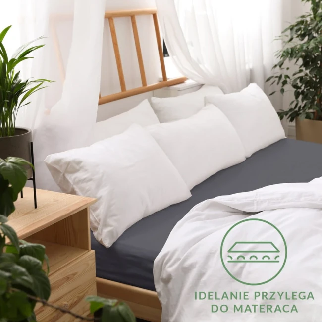 Prześcieradło jersey z gumką w kolorze antracytowym, rozmiar 180x200 cm, rozłożone na łóżku z białymi poduszkami. Widoczne na zdjęciu jest logo w zielonym kolorze oraz tekst IDEALNIE PRZYLEGA DO MATERACA. Tło w jasnej aranżacji pokoju.