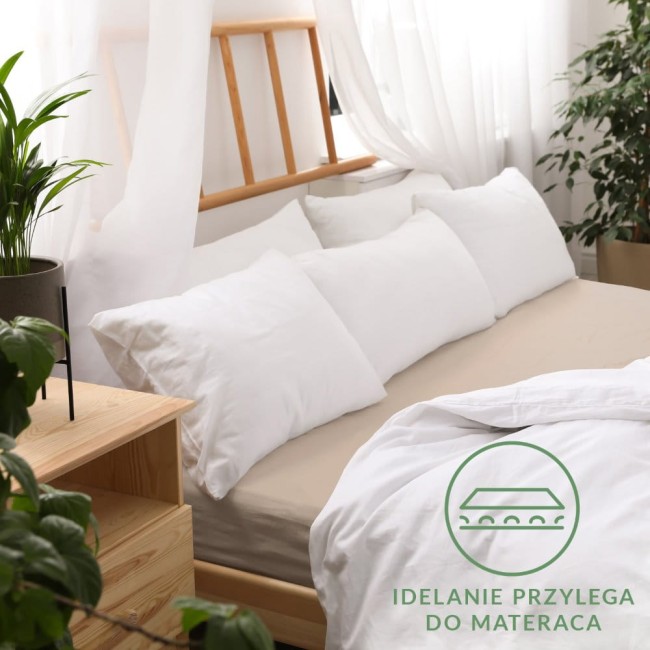 Prześcieradło jersey z gumką 90x200 w kolorze beżowym, rozłożone na łóżku, z widocznym białym zestawem poduszek w tle. Tło z jasnymi zasłonami i rośliną doniczkową. Widoczny napis Idealnie przylega do materaca.