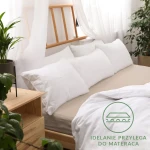 Prześcieradło jersey z gumką w kolorze beżowym, rozmiar 120x200 cm, leżące na łóżku z widoczną narzutą i białymi poduszkami. Tło w jasnych, naturalnych kolorach, z dodatkowymi roślinami doniczkowymi. Widoczne logo w kolorze zielonym w dolnym rogu.