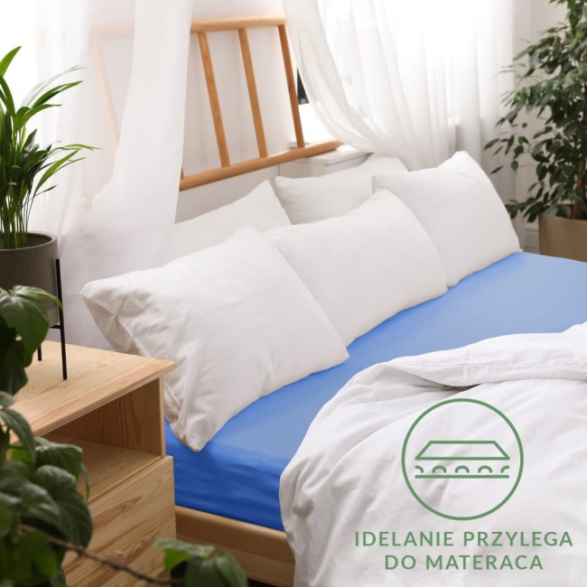 Prześcieradło jersey z gumką 200x220 w kolorze chabrowym, leżące na łóżku z białymi poduszkami. Materiał gładki, dobrze przylegający do materaca. Tło stanowi jasne wnętrze z naturalnymi akcentami roślinnymi. Widoczne detale, jak elastyczna gumka.