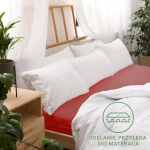 Prześcieradło jersey z gumką 140x200 w kolorze czerwonym, rozłożone na łóżku, z wyraźnym podbiciem krawędzi. W tle białe poduszki i firany, stół nocny z rośliną. Widoczny napis IDEALNIE PRZYLEGA DO MATERACA na białym tle.