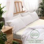 Prześcieradło jersey z gumką w kolorze ecru, rozmiar 90x200, leży na łóżku z białymi poduszkami. Widok z góry, uchwycono cały materac. Na tkaninie widoczna jest tekstura dzianiny. W tle drewniane meble i rośliny, proste tło.