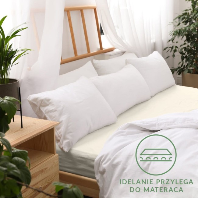 Prześcieradło jersey z gumką w kolorze ecru, o wymiarach 160x200 cm, ułożone na łóżku z białymi poduszkami. Tło jasne, z drewnianym wezgłowiem.
