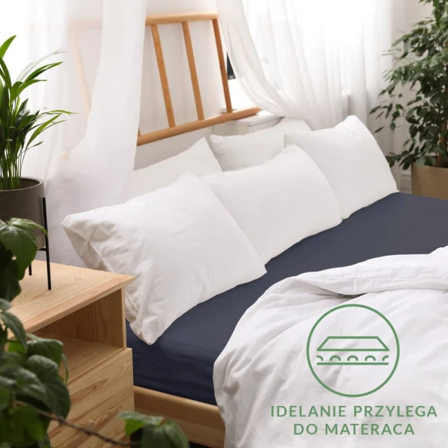 Prześcieradło jersey z gumką 160x200 w kolorze ciemnego granatu, leżące na łóżku. Widoczna gładka struktura materiału, z gumką umożliwiającą dopasowanie do materaca. W tle białe poduszki i osłona łóżka z drewna. W minimalistycznej aranżacji wnętrza.