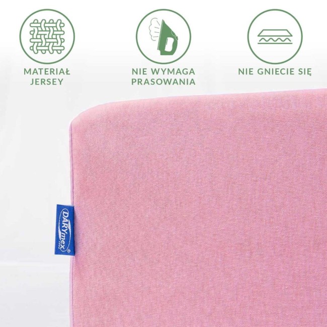 Prześcieradło jersey z gumką w kolorze jasnego różu, widok z boku. Materiał nie wymaga prasowania i jest odporny na gniecenie. Na krawędzi widoczna niebieska metka z logo producenta. Tło białe. Wymiary to 180x200 cm.