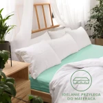 Prześcieradło jersey z gumką 180x200 w kolorze morskim, leżące na łóżku z białą pościelą i poduszkami. Ujęcie z boku, kładąc nacisk na gładką fakturę. Widoczne logo na tle łóżka z drewnianym stelażem, w przestronnym wnętrzu z roślinnością.