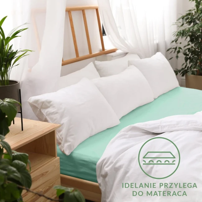 Prześcieradło jersey z gumką w kolorze morskim, rozmiar 90x200, leży na łóżku z białymi poduszkami. Widoczna faktura materiału, idealnie przylega do materaca. W tle roślinność i naturalne światło tworzą przytulną atmosferę.