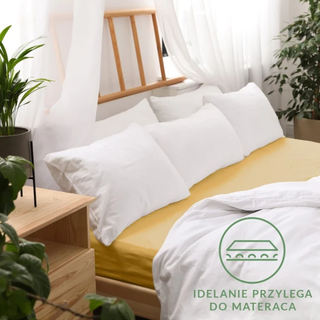 Prześcieradło jersey z gumką 90x200 w kolorze musztardowym, rozłożone na łóżku z białymi poduszkami. W ujęciu z góry, widoczny delikatny materiał. Tło z białymi zasłonami oraz rośliną w doniczce. Logo Idealnie przylega do materaca na dnie zdjęcia.