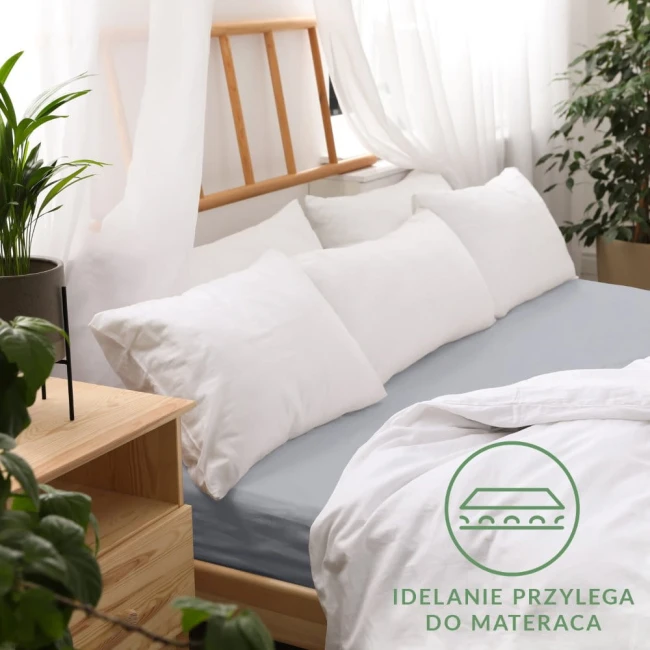 Prześcieradło jersey z gumką w kolorze popielatym, rozmiar 180x200 cm, rozłożone na materacu. W tle widoczne poduszki w białych poszewkach oraz drewniane łóżko, otoczone roślinnością. Na zdjęciu widoczny zestaw białych poduszek.