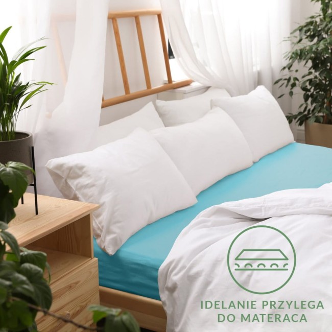 Prześcieradło jersey z gumką 80x160 w kolorze turkusowym, rozłożone na łóżku z białą pościelą. Kąt ujęcia frontalny, z wyraźnym widokiem na elastyczność materiału. Tło jasne z dekoracyjną roślinnością i drewnianymi meblami.