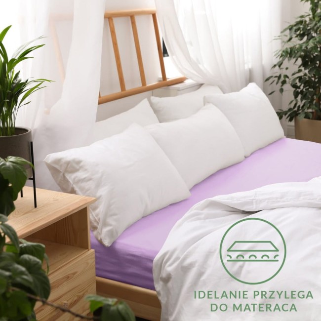 Prześcieradło jersey z gumką 140x200 w kolorze wrzosowym, rozłożone na łóżku z białymi poduszkami. Widoczne gładkie wykończenie materiału. Kontrast w kolorach z jasnym otoczeniem. W dolnym rogu logo i napis „idealnie przylega do materaca”.