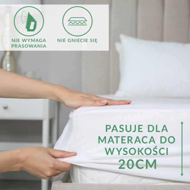 Prześcieradło frotte z gumką w kolorze białym, rozłożone na materacu o wysokości 20 cm. Na zdjęciu widoczne są detale gumki oraz tekst informujący o dopasowaniu do materaca. Kąt ujęcia z boku z ręką trzymającą materiał. Tło neutralne.