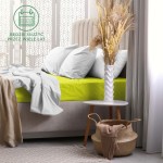 Prześcieradło frotte z gumką 180x200 w kolorze limonka leży na łóżku, otoczone białymi poduszkami. Widoczne jest również białe, okrągłe stolik z kwiatami oraz pleciona kosz z brązowym ręcznikiem. Tło stanowi jasna zasłona i dekoracyjny motyw na ścianie.