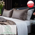 Prześcieradło satynowe z gumką 180x200 w kolorze ciemnego turkusu, leżące na łóżku. Ujęcie frontalne, ukazuje gładką powierzchnię oraz dwa poduszki w odcieniu ciemnego brązu.