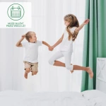 Ochraniacz na materac 180x200 cm w kolorze białym, umieszczony na łóżku. Dzieci skaczą na materacu, co sugeruje jego wygodę i trwałość. Tło stanowią białe zasłony i zielone firany. Widoczny jest również fragment drewnianego wezgłowia.