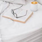 Prześcieradło bawełniane hotelowe białe 140x240 leży na łóżku, na nim otwarta książka z tekstem na stronach oraz okulary. Obok znajduje się filiżanka z kawą na białym tle. Ujęcie z góry, zbliżenie na szczegóły aranżacji.