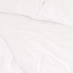 Prześcieradło bawełniane hotelowe w kolorze białym, o wymiarach 140x220 cm, rozłożone na łóżku. Zgładka faktura materiału, brak widocznych wzorów lub dekoracji. Widoczne poduszki w tym samym kolorze na tle jasnej pościeli.