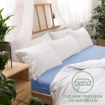 Prześcieradło jersey z gumką w kolorze niebieskim, rozmiar 140x200, rozłożone na łóżku. Na zdjęciu widoczna jest tekstura materiału, złożone białe poduszki w tle oraz elementy drewnianego łóżka. Tło jest jasne, w aranżacji pokojowej. Logo na tkaninie.