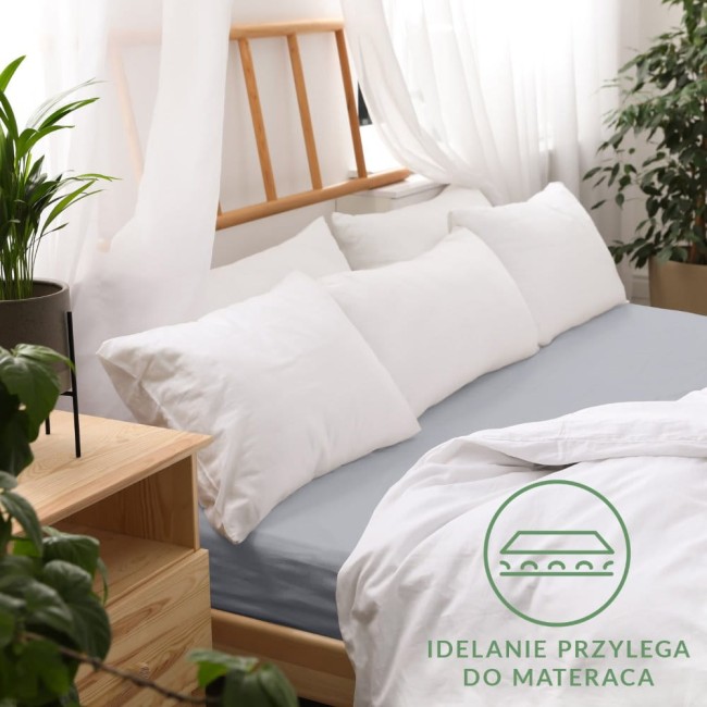 Prześcieradło jersey z gumką 140x200 w kolorze popielatym, rozłożone na łóżku z białymi poduszkami w tle. Materiał gładki, przyjemny w dotyku. W widocznym rogu znajduje się napis IDEALNIE PRZYLEGA DO MATERACA. W otoczeniu roślinność i drewniane meble.