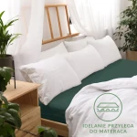 Prześcieradło jersey z gumką o wymiarach 140x200 w kolorze zieleni butelkowej. Leży na łóżku z białymi poduszkami i narzutą, dobrze przylegając do materaca. W tle widoczne akcenty roślinne na drewnianej komodzie oraz komplet poduszek.
