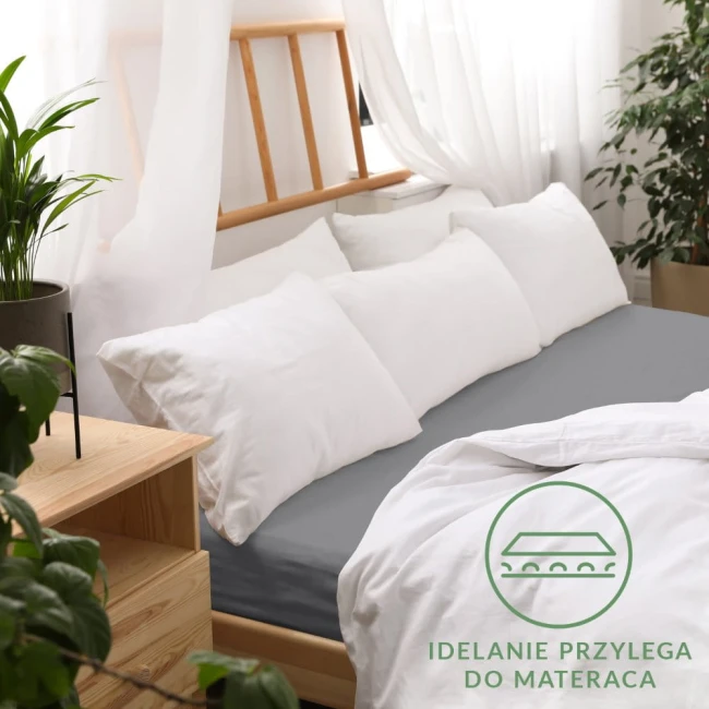 Prześcieradło jersey z gumką 180x200 w kolorze szarym, leżące na łóżku z białymi poduszkami. Dobry uchwyt do materaca, tekstura gładka. Tło to jasne zasłony i drewniane meble, dodatkowo roślina doniczkowa. Widoczny napis IDEALNIE PRZYLEGA DO MATERACA.