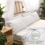 Prześcieradło jersey z gumką 140x200 w kolorze ecru, leżące na łóżku z białymi poduszkami. Widoczne wykończenie gumką, idealnie przylegające do materaca. Tło z drewnianym zagłówkiem, rośliną i minimalistycznym wystrojem.