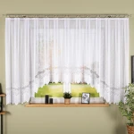 Firana gotowa MODESTA żakardowa, biała, 500x160 cm, wisząca na karniszu. Charakteryzuje się delikatnym, żakardowym wzorem oraz falbaniastym wykończeniem na dole. Tło jasne, elementy dekoracyjne w postaci roślin doniczkowych i zdjęcia na parapecie.
