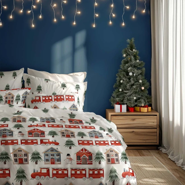 Pościel bawełniana 220x200 w czerwony wzór z pociągiem, zimowymi domkami i choinkami. Widoczna na łóżku z poduszkami w tym samym wzorze, otoczona dekoracjami świątecznymi oraz choinką w tle. Tło w ciemnoniebieskim kolorze.