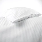 Poszewka na poduszkę 40x40 w kolorze białym z drobnymi paskami o szerokości 1 cm, leżąca na gładkiej powierzchni. Zapięcie na zakładkę, gładka faktura tkaniny, bez widocznych napisów czy logo. Ujęcie z boku, pokazujące dwa złożone rogi.