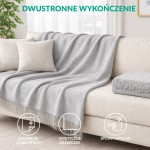 Koc bawełniany 200x220 cm w szarym kolorze, z frędzlami na jednym końcu, położony na jasnym fotelu. Tło stanowi jasne okno z roletami i delikatne, minimalistyczne elementy dekoracyjne. Koc sprawia wrażenie miękkiego i przytulnego.