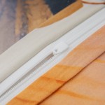 Zbliżenie na zamek błyskawiczny poszewki pościeli, z białym suwakiem i osłoną w kolorze kremowym. Widoczna tekstura materiału oraz delikatne zabarwienie w odcieniach beżu i pomarańczy. Poszewka leży na gładkiej powierzchni.
