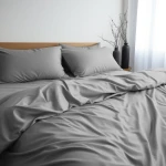 Poszewka satynowa 70x80 w kolorze popielatym, leżąca na łóżku. Zestaw składa się z dwóch poduszek i poszewki na kołdrę, widoczne na tle jasnych zasłon. Materiał gładki, błyszczący, podkreślający elegancję.