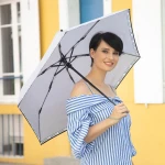 Parasol mini w kolorze białym z czarnym paskiem. Złożony, trzymany przez osobę w pasiastym topie. Widoczna struktura czaszy i uchwyt. Tło w jasnych kolorach, na złocistym murze budynku. Idealny do ochrony przed deszczem.